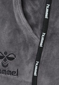 Grauer Stoff mit einer weichen Textur, der ein schwarzes gesticktes Logo und einen schwarzen Kordelzug mit "hummel" in weißem Text aufweist.