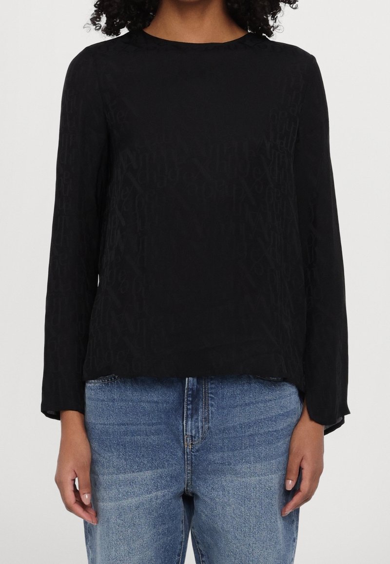 Femme portant un chemisier noir à manches longues avec une texture subtile et un jean bleu en denim, debout devant un fond clair uni.