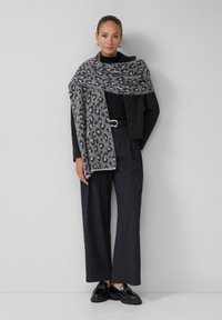 Écharpe à imprimé léopard gris drapée sur une veste noire courte, associée à un pantalon à rayures verticales et des chaussures noires brillantes.