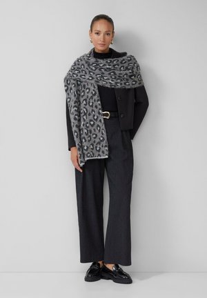 Écharpe à imprimé léopard gris drapée sur une veste noire courte, associée à un pantalon à rayures verticales et des chaussures noires brillantes.