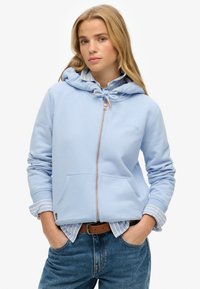 Sudadera con capucha azul claro con cremallera, capucha con cordones, bolsillos delanteros y puños acanalados, combinada con una camisa de rayas con cuello y jeans de mezclilla.