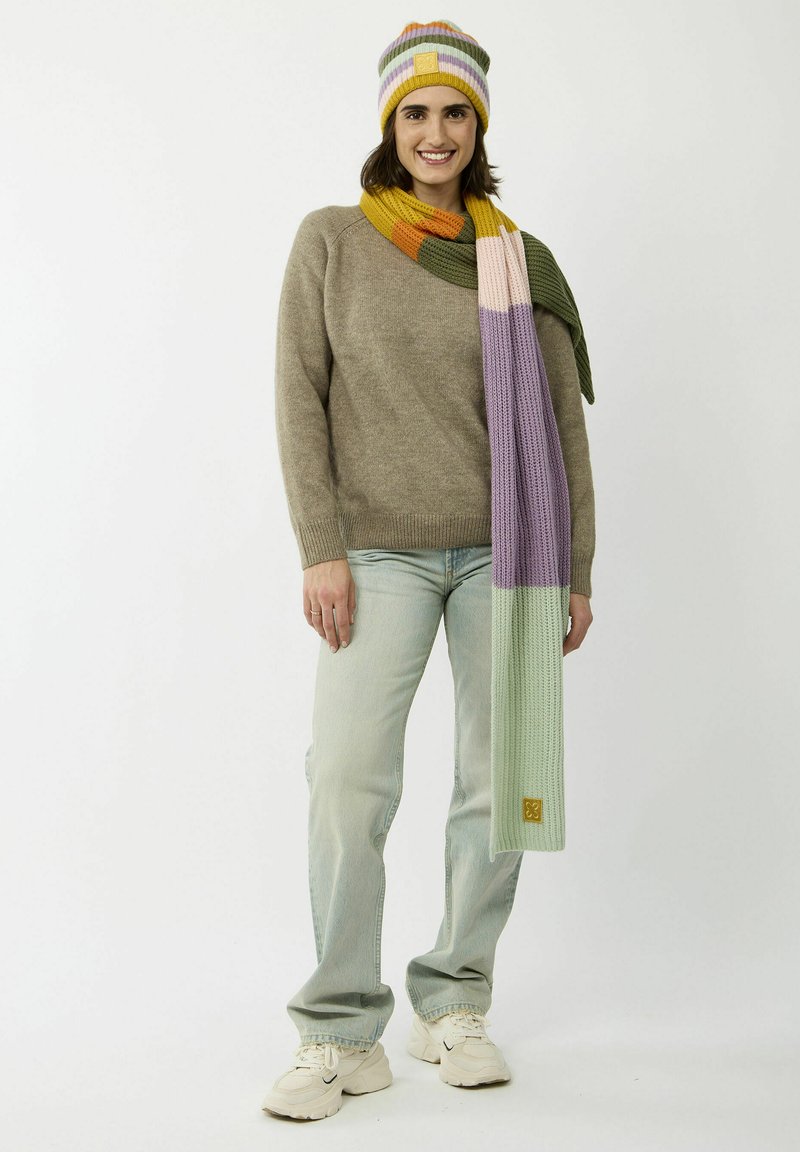 Gehaakte beige trui, meerkleurige gestreepte hoed en sjaal, lichtblauwe jeans en witte sneakers. De sjaal heeft een gladde textuur met verloopkleuren.