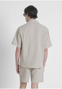 Camicia beige a maniche corte con una trama testurizzata e stropicciata. Abbinata a pantaloni corti beige coordinati, con cuciture visibili e una vestibilità rilassata.