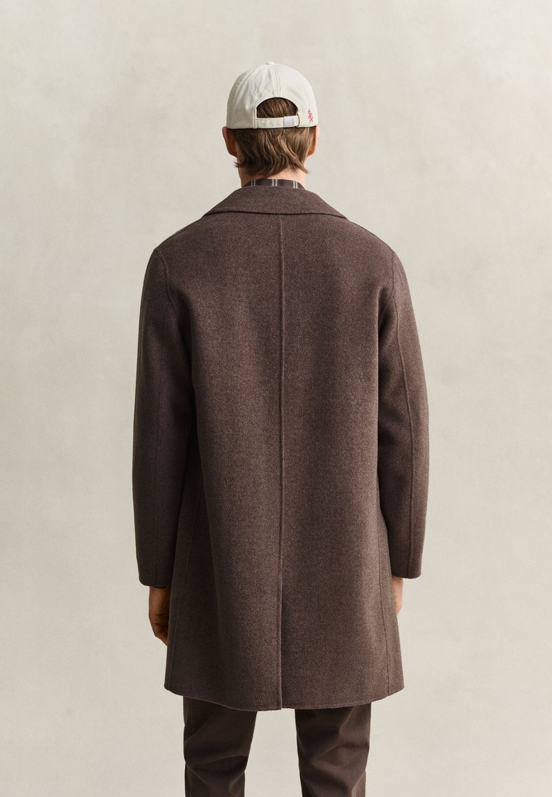 Uomo che indossa un lungo cappotto marrone, pantaloni scuri e un berretto da baseball bianco, visto di spalle davanti a uno sfondo chiaro e semplice.