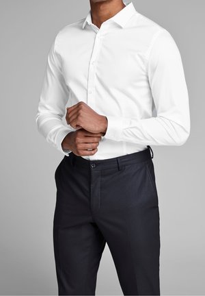 Homme portant une chemise blanche cintrée et un pantalon noir formel, ajustant la manche gauche de sa chemise sur un fond gris uni.