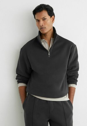 Mann mit dunklen Haaren, trägt ein schwarzes Sweatshirt mit halbem Reißverschluss und passende Hose, steht mit den Händen in den Taschen vor einer weißen, strukturierten Wand.