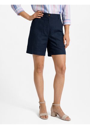 Vrouw draagt marineblauwe shorts met hoge taille, gestreept shirt met lange mouwen en beige sandalen met blokhak en enkelriempje, staat met één hand op haar heup.