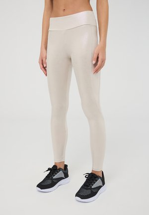 Leggings beige e lucidi con una cintura larga e liscia. Realizzati in materiale elasticizzato. Abbinati a scarpe da ginnastica nere con accenti grigi.
