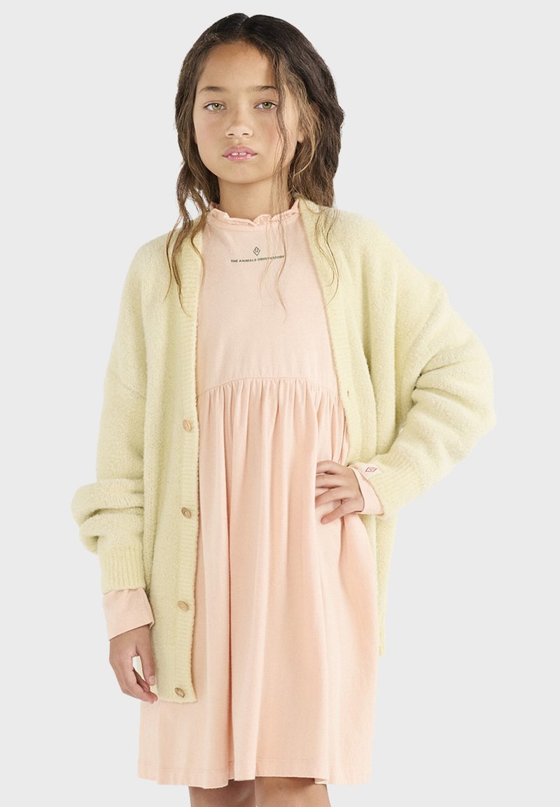 Hellgelbe Strickjacke mit Knöpfen, getragen über einem rosa, kurzärmeligen Kleid mit geraffter Taille und Rüschenausschnitt.