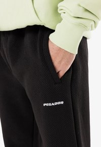 Hand in der Tasche, trägt schwarze Pegador-Hosen und ein hellgrünes langärmliges Hemd oder Sweatshirt.