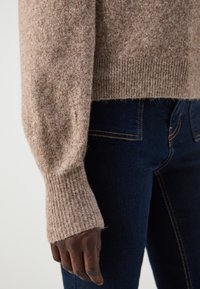 Lys brun, teksturert strikkegenser med ribbestrikkede mansjetter, lagd over mørke denim jeans, som fremhever en avslappet stil og varmt materiale.