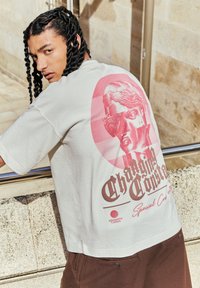 White oversized t-shirt featuring a pink graphic of a statue and text, with short sleeves and a relaxed fit. Casual brown shorts worn.