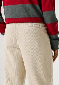 Beige korduroja bikses ar vertikālām ribām un aizmugurējām kabatām. Apvienotas ar sarkanu un pelēku svītrainu garo piedurkņu topu.