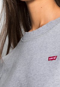 Sudadera gris hecha de una mezcla de algodón, con cuello redondo acanalado y un parche del logo de Levi's en rojo en el pecho. Textura suave visible.