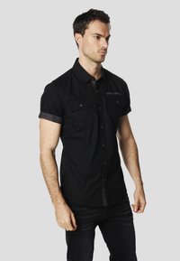 Camisa negra de manga corta con botones, dos bolsillos frontales, mangas remangadas y un detalle sutil del logo cerca del escote. Diseño de ajuste casual.
