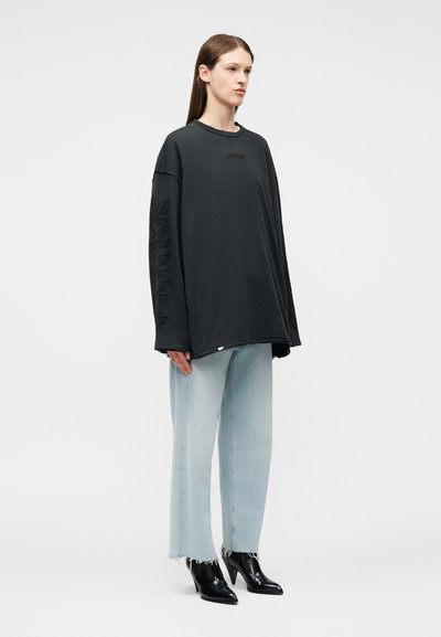 Γυναίκα που στέκεται φορώντας μια φαρδιά μαύρη φούτερ μπλούζα με κεντημένα μανίκια, ανοιχτό γαλάζιο τζιν παντελόνι με φαρδιά μπατζάκια και μαύρα μυτερά μποτάκια με τακούνι.