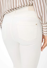 Weiße, schmale Jeans mit Gesäßtaschen, aus Denim. Mit einem kleinen goldenen Akzent an der rechten Tasche. Texturiert mit einer glatten Oberfläche.