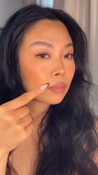 Primo piano di una persona che indica la propria guancia. Ha la pelle di un marrone medio, capelli neri lunghi e mossi, e indossa un trucco neutro con una finitura opaca.