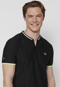 Juodas polo marškinėliai su tekstūruotu raštu, turintys dviejų mygtukų užsegimą, žaliu apvadu ant apykaklės ir rankovių, bei logotipu ant krūtinės.