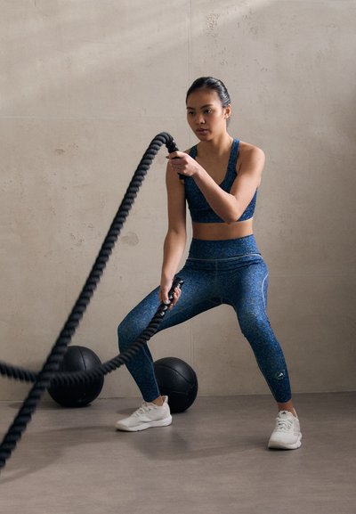 Conjunto atlético de patrón azul con un sujetador deportivo y leggings de cintura alta. Cuerda de batalla oscura en uso, zapatos deportivos blancos y balones medicinales negros cerca.