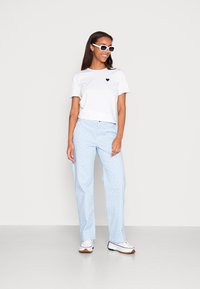 T-shirt curta branca com um pequeno logótipo de coração preto, combinada com calças xadrez azuis e sapatilhas brancas com pormenores castanhos.