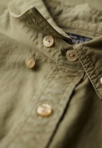 Camisa abotonada de color verde oliva con un tejido suave y texturizado, un cuello y botones de color claro. Costuras y colocación de botones notable.