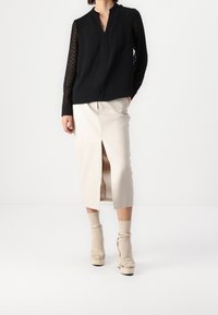 Nainen pukeutunut mustaan pitkähihaiseen paitaan, jossa on läpinäkyvät sahalaitahihat, vaaleanbeigeen midi-hameeseen, jossa on etuaukko, vaaleanbeigeihin sukkiin ja vaaleanbeigeihin korokepohjaisiin korkokenkiin.