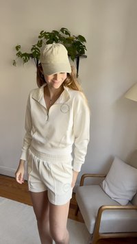 Light beige zip-up sweatshirt with funnel neck, matching shorts featuring a logo patch. Soft fabric, relaxed fit, visible stitching details.