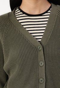 Olivovo zelený pletený kardigán s výstrihom do V, piatimi gombíkmi a rebrovanou textúrou, prehodený cez pásikové tričko v čiernej a bielej farbe.