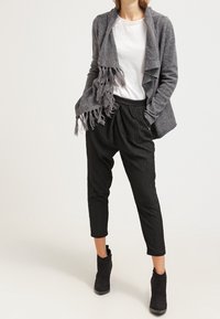 Šedý kardigan s třásněmi přes bílé tričko, doplněný černými ribbed tapered kalhotami a černými kotníkovými botami.