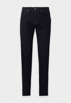Homme debout, une main dans la poche, portant un polo beige à manches courtes, un pantalon slim foncé, des chaussettes blanches et des baskets beiges sur fond blanc.