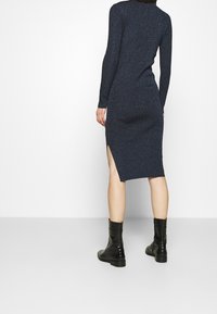 Abito lungo a maniche con design aderente di colore blu navy, con spacco laterale e texture luminosa, abbinato a stivaletti neri alla caviglia.