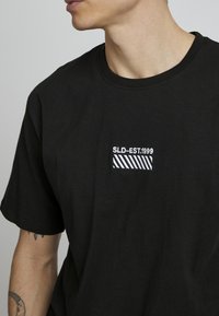 Musta puuvillainen T-paita pyöreällä kaula-aukolla, jossa on pieni valkoinen brodeeraus, joka lukee "SLD-EST.1999" sekä vinoraitoja.