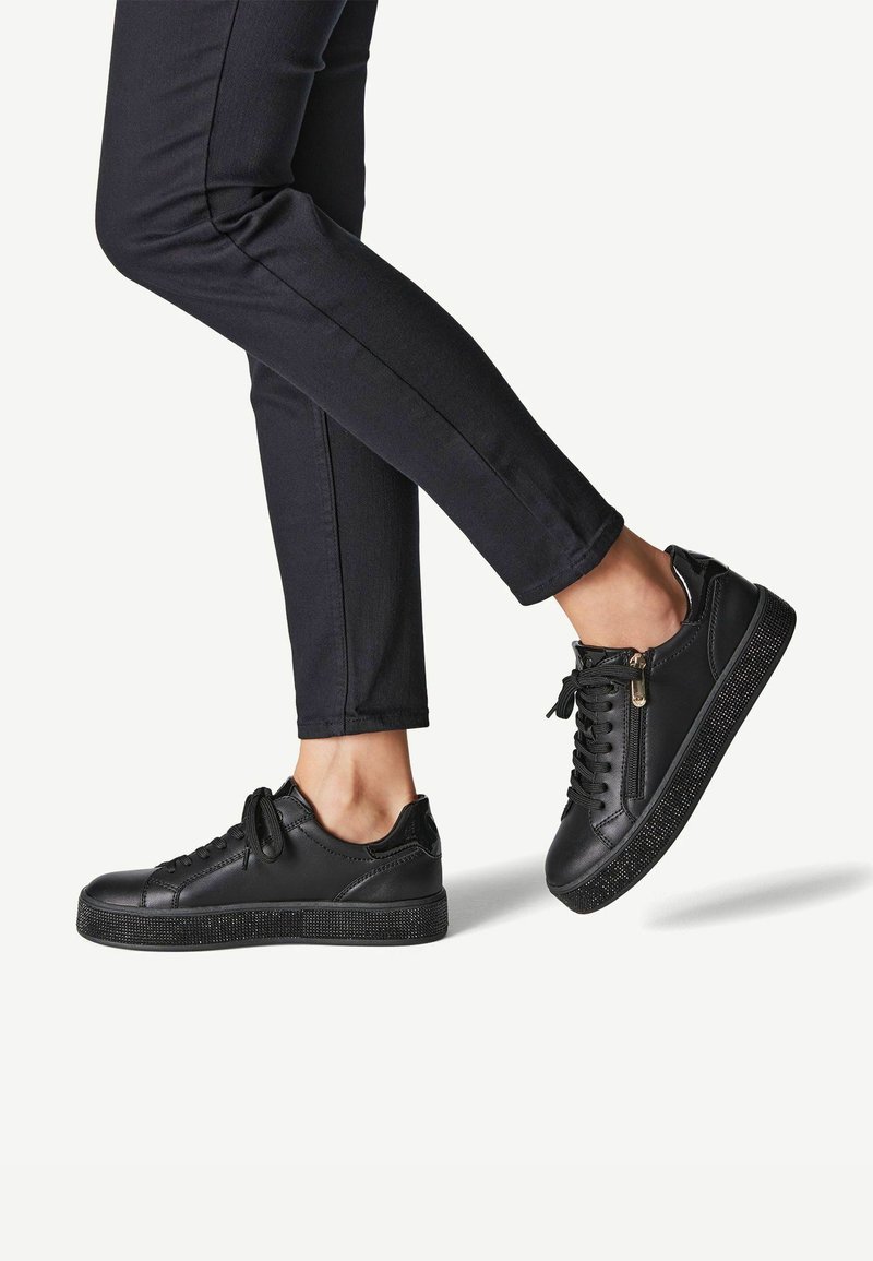 Sneaker in pelle nera con suola platform nera testurizzata, caratterizzate da dettagli in zip dorati e lacci abbinati, indossate con pantaloni slim-fit scuri.