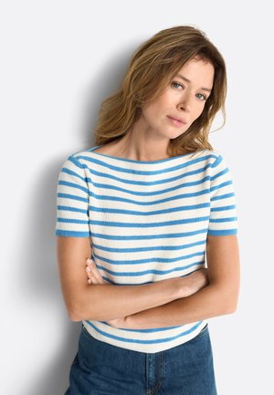 Donna con capelli castano chiaro in piedi con le braccia incrociate, indossa una maglietta a maniche corte bianca e blu a righe e jeans blu su sfondo bianco.