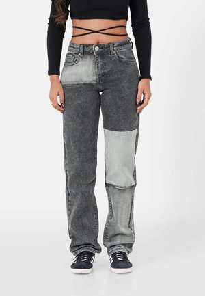 Vrouw draagt hooggetailleerde, tweekleurige zwart-grijze patchwork jeans, gecombineerd met zwarte vetersneakers en een zwarte cropped top met wikkelbanden.