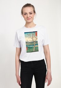 Bílé bavlněné tričko s barevným potiskem krajiny se stromem, vodou a horami, doplněné červeným textem na designu.