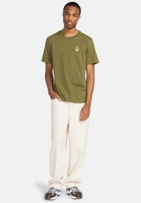 Camiseta de algodón verde oliva con mangas cortas y un pequeño logo gráfico. Combinada con pantalones de corte suelto color crema y zapatillas.