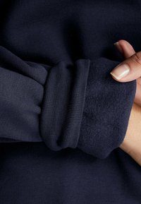 Detail na manžetě námořnické modré mikiny, s texturovaným ribovým pletením a měkkým fleece povrchem uvnitř, s hladkým vnějším povrchem.