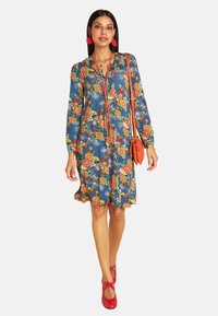 Blumenkleid in Blau mit orangefarbenen und weißen Mustern, langen Ärmeln, V-Ausschnitt und ausgestelltem Saum. Kombiniert mit roten Schuhen und Accessoires.