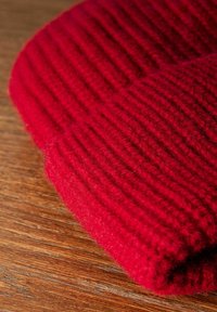 Bonnet en laine rouge tricoté avec une texture côtelée, montrant des points détaillés et une apparence douce et duveteuse, reposant sur une surface en bois.
