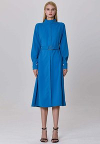 Robe longue bleu à manches longues avec un col montant, taille ceinturée avec une boucle circulaire, et une jupe évasée fluide, fabriquée en tissu lisse.