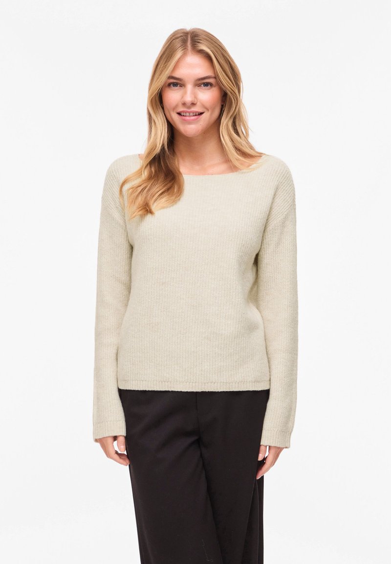 Πλεκτό πουλόβερ σε ανοιχτό μπεζ χρώμα με στρογγυλή λαιμόκοψη, μακριά μανίκια και χαλαρή εφαρμογή, συνδυασμένο με σκούρο παντελόνι. Ορατές λεπτομέρειες υφής στο ύφασμα.