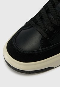 Tenis cu un amestec de piele netedă și suede texturat, bot rotund, șireturi negre și o talpă din cauciuc alb contrastant.