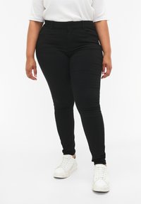 Črne skinny jeans s srednje visokim pasom, petimi žepek in gladkim, raztegljivim materialom. V kombinaciji z belimi športnimi copati.
