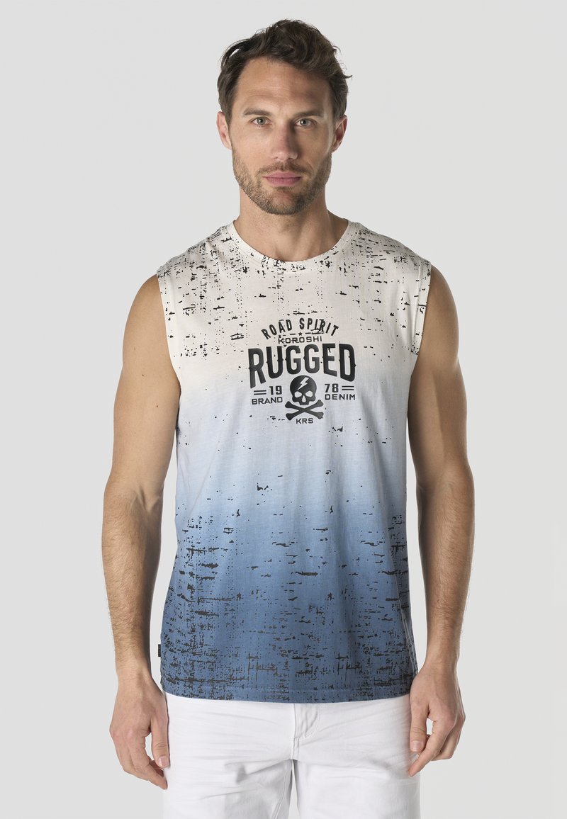 Hombre con cabello corto y castaño que lleva una camiseta sin mangas blanca con degradado azul, con el texto "Rugged" y un gráfico de calavera, de pie frente a un fondo liso.
