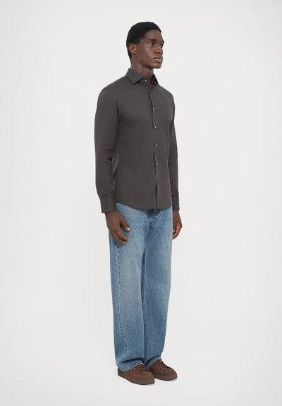 Mørkegrå langermet skjorte, med knappelukking, sammen med lyseblå vide jeans og brune joggesko, stående foran en ensfarget bakgrunn.