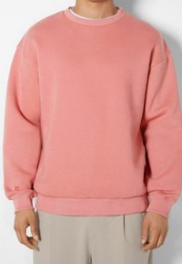 Person som bär en enkel rosa crewneck-tröja och ljusbeiga byxor, stående framför en vit bakgrund.