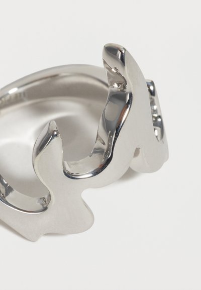 Λαμπερό ασημένιο δαχτυλίδι με αφηρημένο, κυματιστό σχέδιο που σχηματίζει δύο καμπύλες προς τα πάνω, αντανακλώντας το φως σε απλό λευκό φόντο.