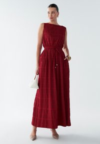 Rød maxikjole med tekstureret mønster, ærmeløs design og elastisk talje. Kombineret med en hvid håndtaske og pyntede nøgenfarvede hæle.